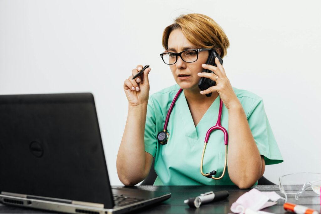 Doctor talking on the phone to a patient while sitting at their desk in front of a laptop. hausoflife.com